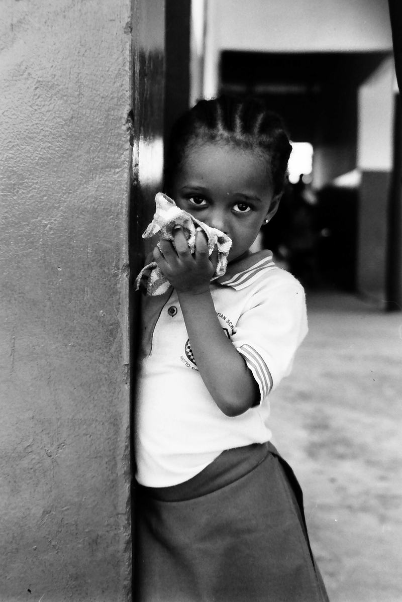 Black-and-white photograph of a young child standing partially behind a wall at a school in Kpandai, Ghana.